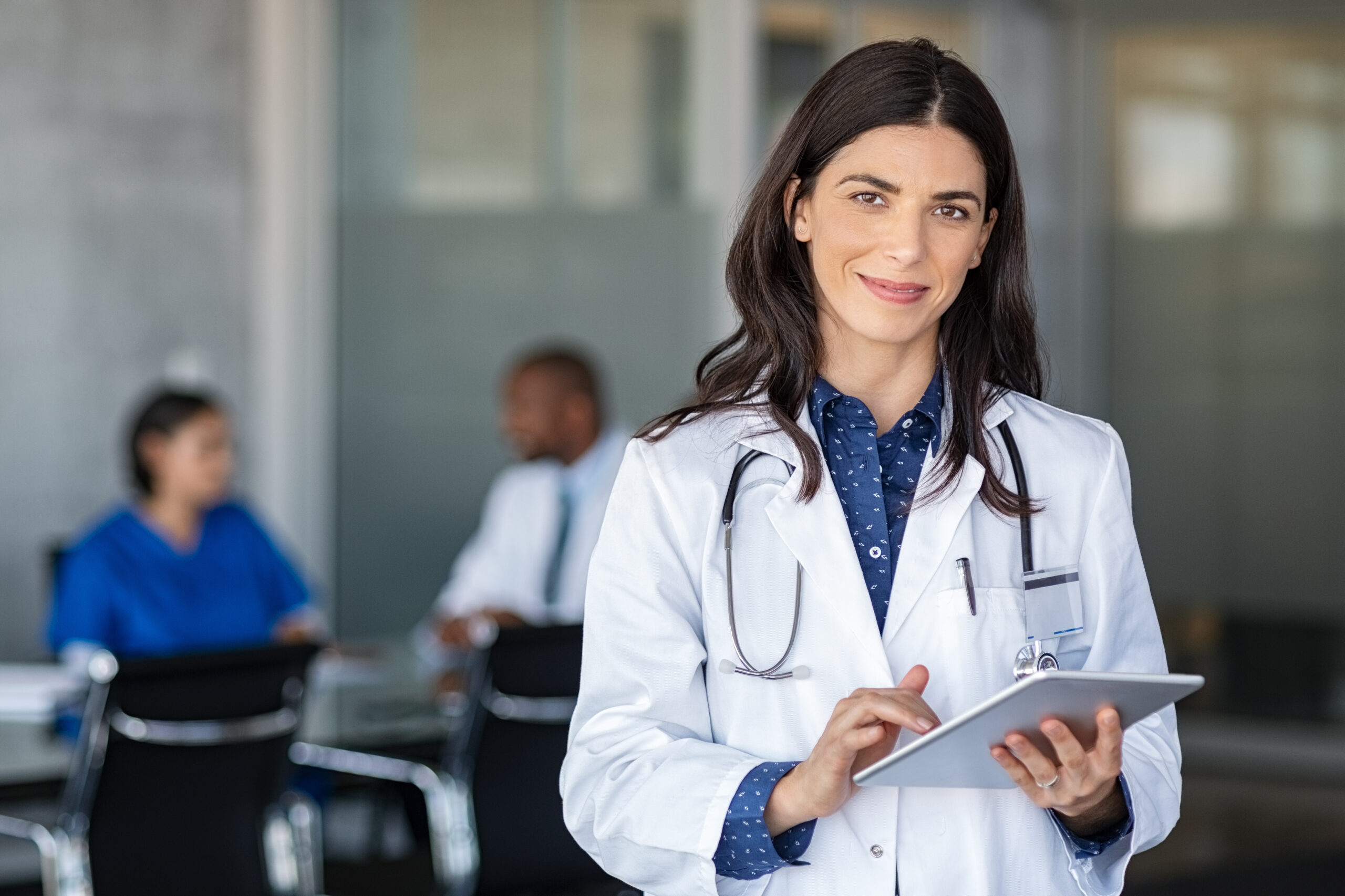 Portrait of beautiful mature woman doctor holding digital tablet and looking at camera. Confident female doctor using digital tablet with colleague talking in background at hospital. Latin nurse in labcoat and stethoscope in private clinic with medical team working.